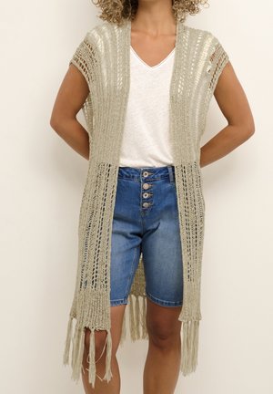 Femme portant un gilet sans manches en maille beige ajourée avec franges par-dessus un t-shirt blanc à col en V et un short en jean bleu à boutons.