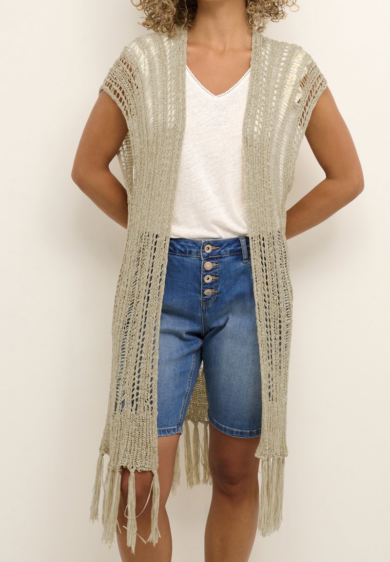 Femme portant un gilet sans manches en maille beige ajourée avec franges par-dessus un t-shirt blanc à col en V et un short en jean bleu à boutons.
