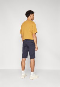 Maglietta corta a maniche per uomo color senape abbinata a pantaloni navy. Sneakers e calze bianche completano il look. Design semplice, senza pattern notevoli.