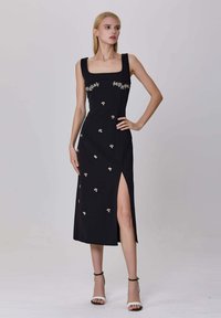 Femme dans une robe midi sans manches noire avec broderies florales et une fente haute, portant des talons à lanières noirs et blancs, se tenant devant un fond uni.