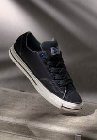 Black canvas sneaker with blue eyelets, white rubber sole, and textured toe cap. Features contrast stitching and an "All Star" logo patch.