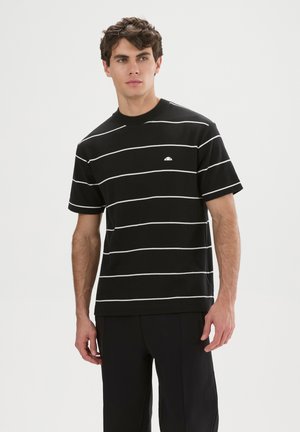 Junger Mann trägt schwarzes Kurzarmshirt mit weißen horizontalen Streifen und schwarze Hose, steht vor einfachem hellem Hintergrund.