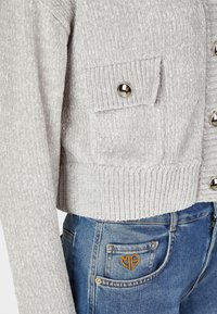 Hellgraue, taillierte Jacke aus strukturiertem Stoff mit Metallknöpfen und einer Brusttasche. Kombiniert mit blauen Jeans.