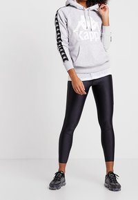 Felpa grigia con logo Kappa bianco, strisce laterali nere con motivo del logo, abbinata a leggings neri e sneaker neri. Texture liscia.