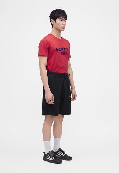 Jeune homme debout face à l'avant portant un t-shirt rouge Dsquared2 Milano, un short noir, des chaussettes blanches et des baskets noires Adidas.
