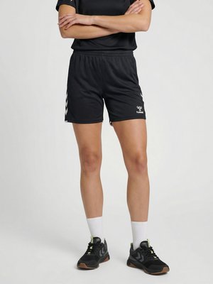Person trägt schwarze Sportshorts mit weißen Chevron-Akzenten, schwarze Sneaker, weiße Socken und ein schwarzes Shirt mit verschränkten Armen.