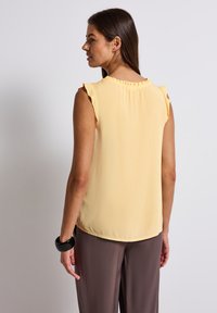 Blouse sans manches jaune avec un col froncé, texture douce et coupe ample, assortie à un pantalon marron et un bracelet noir.