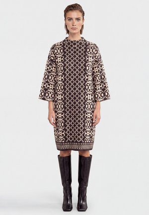 Woman wearing a knee-length brown dress with beige geometric patterns and wide sleeves, paired with tall black leather boots.