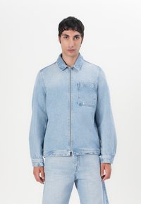 Lys blå denimjakke med frontlukking, spiss krage og en enkelt brystlomme. Har sydd detaljer og lange ermer.