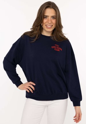 Junge Frau trägt einen marineblauen Pullover mit rotem Schriftzug "Feel Good Club" und weiße Hosen, lächelt mit der Hand in der Hüfte.