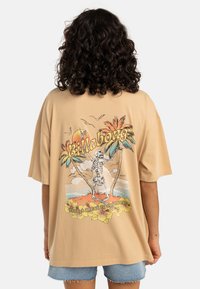 Beige oversized t-skjorte med et grafikk av et skelet mellom palmer, med solnedgang og blomsterdetaljer nederst.