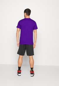 T-shirt en coton violet, manches courtes, vue de dos, associé à un short noir, baskets rouges et blanches, design minimaliste, sans motifs visibles.