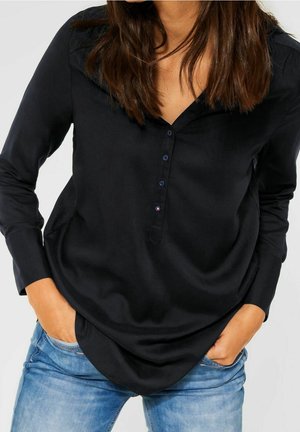 Kvinde iført en sort langærmet skjorte med knapper, hænderne i lommerne på lyseblå jeans, stående mod en hvid baggrund.