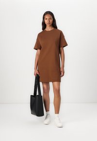 Bruine katoenen t-shirt jurk met korte mouwen en een relaxte pasvorm, gecombineerd met een zwarte tote tas en witte sneakers. Gladde textuur overal.