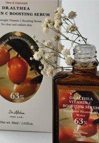 Botella de vidrio ámbar con forma cuadrada y tapa texturizada, con etiqueta de producto que presenta gráficos en naranja y verde, colocada al lado de flores blancas.