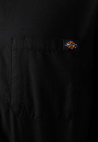 Black short-sleeve shirt with a front pocket. Features a square label displaying the "Dickies" logo. Made of a smooth, durable fabric.
