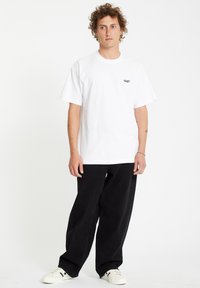 Weißes Baumwoll-T-Shirt mit kurzen Ärmeln, das ein kleines schwarzes Grafikdesign auf der linken Brust zeigt. Kombiniert mit schwarzen, locker sitzenden Hosen und Sneakers.
