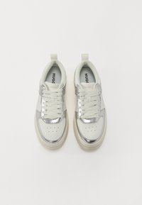 Baskets à lacets avec un embout en métal argenté, tige en cuir blanc et semelle beige texturée. Marques visibles sur la languette.