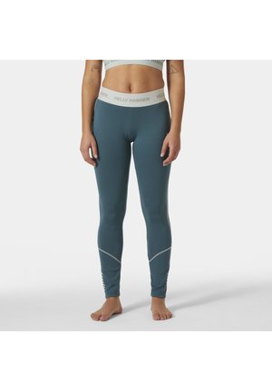 Mujer con leggings ajustados en color verde azulado y sujetador deportivo a juego con el logo "Helly Hansen", de pie descalza contra un fondo liso.