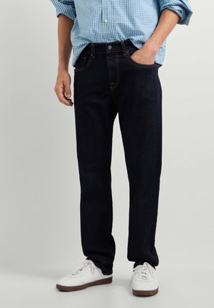 Man draagt donkerblauwe rechte jeans, witte sneakers en een lichtblauw geruit overhemd met één hand in de zak.