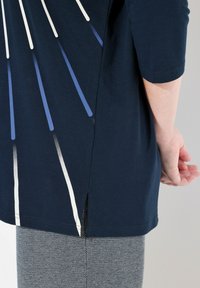 Mörkblå kortärmad topp med geometrisk design som har vita och blå linjer; inkluderar en sidofick. Bärs med grå mönstrade byxor.