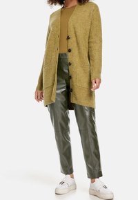 Hořčicově žlutý dlouhý cardigan s otevřeným předním dílem, texturovaný pletený, se dvěma předními kapsami, spárovaný se zelenými koženými kalhotami a bílými teniskami.