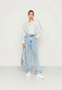 Top de mangas longas azul claro com padrão e mangas bufantes, calças de ganga largas de cintura alta em denim claro e ténis brancos com plataforma. A portar uma mala azul clara.