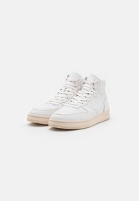 Un par de zapatillas altas blancas con detalles perforados, cordones blancos y suelas de goma beige sobre un fondo blanco liso.