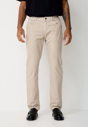 Man wears beige slim-fit trousers, black shoes, and a black shirt, standing against a plain white background.