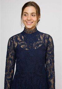 Námornícka čipkovaná sukňa s vysokým golierom, dlhými rukávmi a zložitými kvetinovými vzormi. Priehľadná textúra kontrastuje s pevnou spodnou vrstvou.