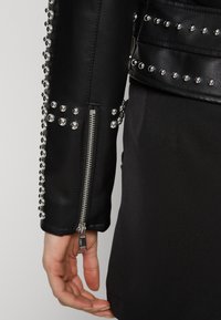 Manche de veste en cuir noir avec des clous argentés le long de la couture et une fermeture éclair. La finition texturée contraste avec le tissu lisse en dessous.