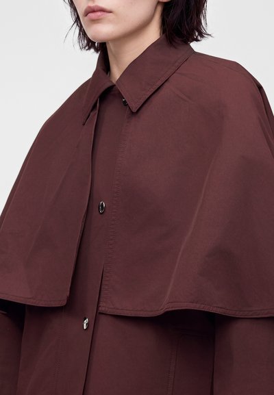 Femme portant une cape manteau oversize boutonnée marron foncé avec un large col, sur un fond clair uni.