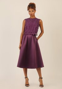 Abito viola con top in pizzo con motivi intricati, gonna in satin A-line liscia e orlo fino al ginocchio. Sandali neri con cinturini.