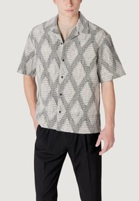 Camicia a maniche corte con bottoni di un beige chiaro con un motivo geometrico nero. Presenta un colletto alla camicia e una vestibilità rilassata. Abbinata a pantaloni neri.