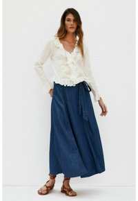 Weiße, gefrontete Bluse mit floralen Details, kombiniert mit einem langen, dunkelblauen Jeanrock mit einem Bindeband in der Taille und braunen Sandalen.
