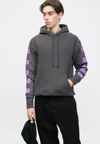 Grauer Kapuzenpullover mit Fronttasche, verziert mit lila grafischen Designs an den Ärmeln. Das Modell trägt schwarze Hosen und hält eine schwarze Mütze.