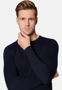 Granatowy sweter z długim rękawem o prążkowanej fakturze, dopasowanym kroju i okrągłym dekolcie; posiada małe logo po lewej stronie.