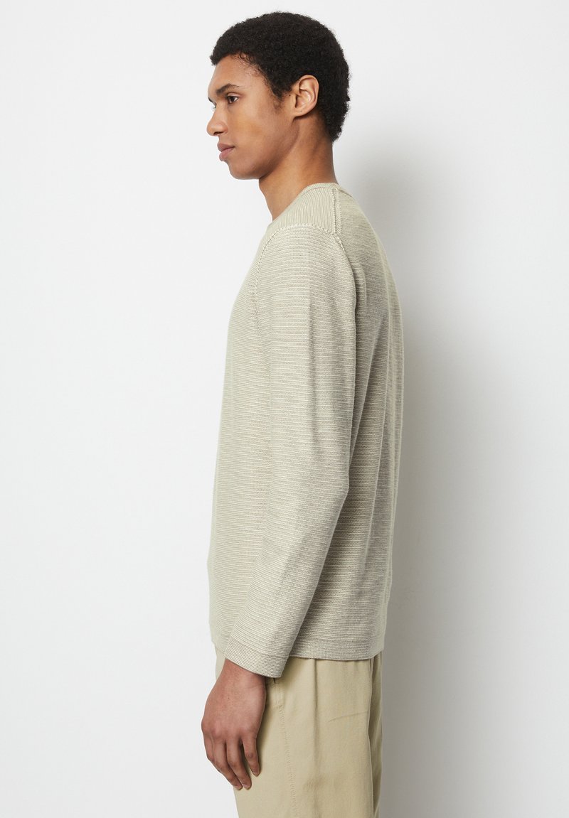 Beżowy sweter dzianinowy z długim rękawem, z teksturowanymi paskami, okrągłym dekoltem i ściągaczami na rękawach, noszony z jasnymi spodniami.