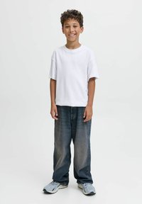Garçon souriant avec des cheveux bouclés, portant un t-shirt blanc, un jean sombre ample et des baskets gris clair, se tenant devant un fond clair uniforme.
