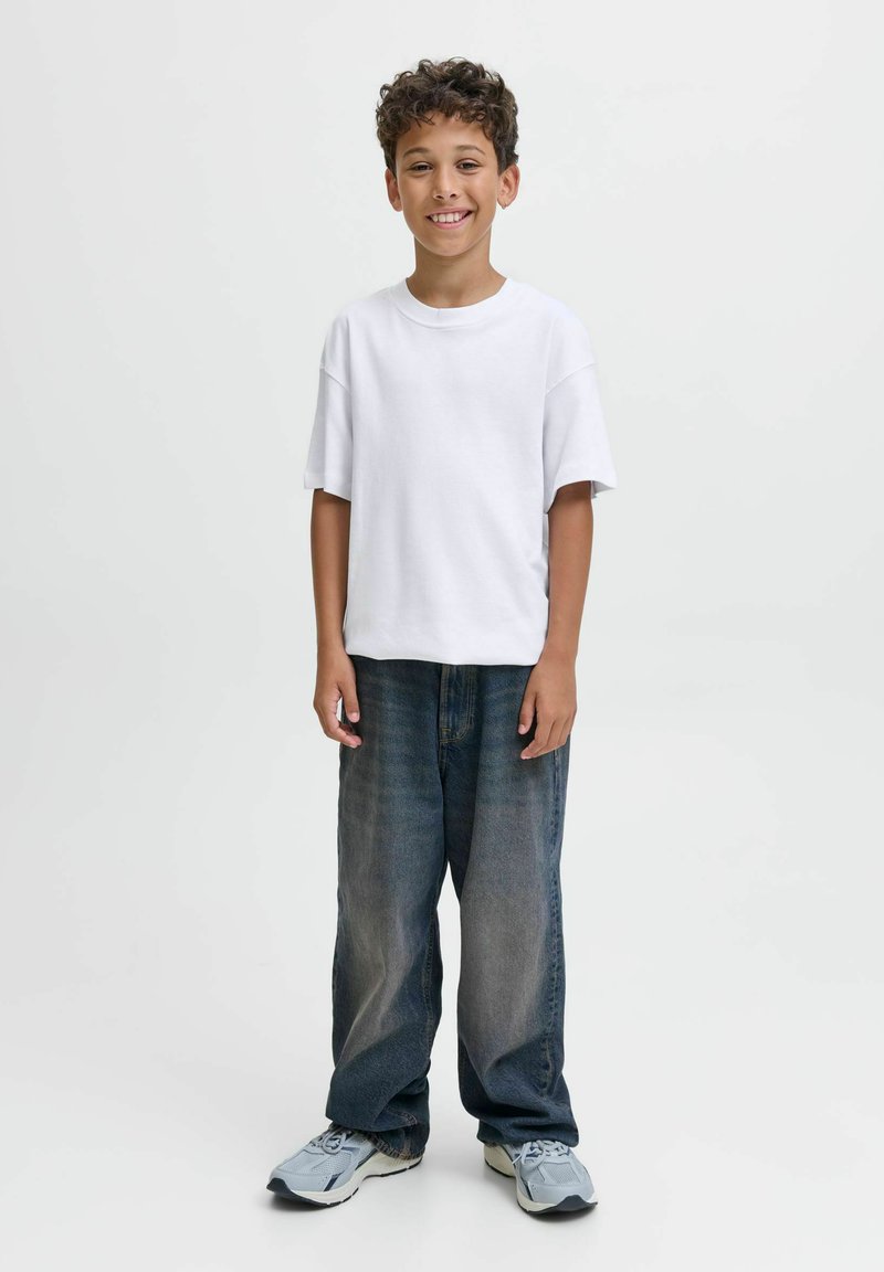 Garçon souriant avec des cheveux bouclés, portant un t-shirt blanc, un jean sombre ample et des baskets gris clair, se tenant devant un fond clair uniforme.