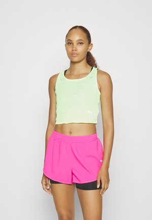 Jeune femme avec des cheveux bouclés en queue de cheval, portant un crop top jaune fluo et un short de sport rose vif sur un fond clair.