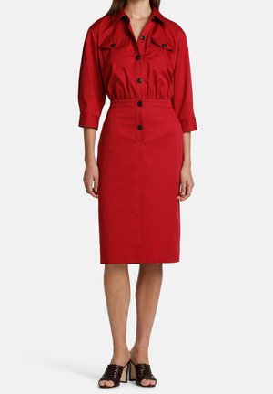 Femme portant une robe rouge boutonnée avec des manches trois-quarts et des poches avant, associée à des mules à talons ouvertes marron foncé.