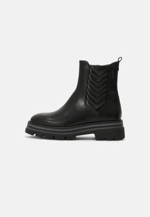 Bottine noire en cuir avec motif chevron matelassé sur les côtés, semelle épaisse et languette à l'arrière, vue de profil sur fond blanc.