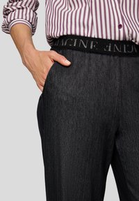 Person mit der Hand in der Tasche einer dunklen Nadelstreifenhose, trägt ein lila-weiß gestreiftes Hemd, das eingesteckt ist, und zeigt einen schwarzen Bund mit Text.