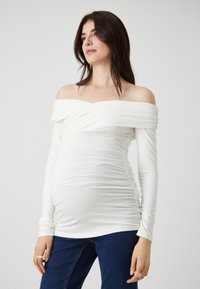 Balta of-of-shoulder palaidinė su ilgomis rankovėmis, susiraukšlėta tekstūra juosmens srityje ir prigludusi išvaizda, derinama su tamsiai mėlynomis džinsomis.