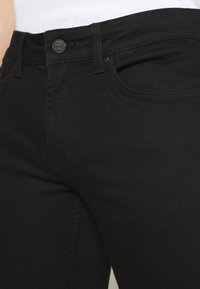 Jeans de mezclilla negra con un corte ajustado, diseño estándar de cinco bolsillos y un botón metálico circular en la cintura con costuras a tono.