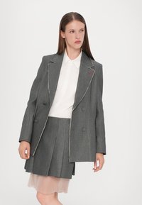 Blazer gris à double boutonnage avec des coutures blanches, associé à une jupe plissée rose. Chemise blanche à boutons en dessous.
