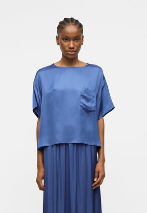 Frau mit geflochtenem Haar, die ein weites, kurzärmeliges, blaues Satintop mit Brusttasche und passendem blauem Rock vor einem schlichten Hintergrund trägt.