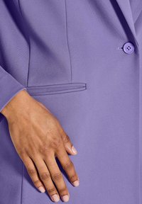 Hand rustend op een lila blazerzak, met zichtbare knoop, naden en een gladde stoftextuur.