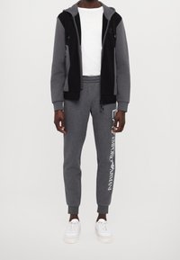 Svart och grå zip-up huvtröja med sidofickor, grå sweatpants med vitt logotryck och vita sneakers. Slät tyg, avslappnad passform.